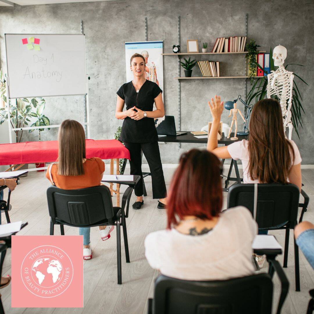 Accredited Beauty Training Courses, a female lecturer in a black beauty therapist outfit, is teaching a class. A therapy bed and skeleton are to either side of her